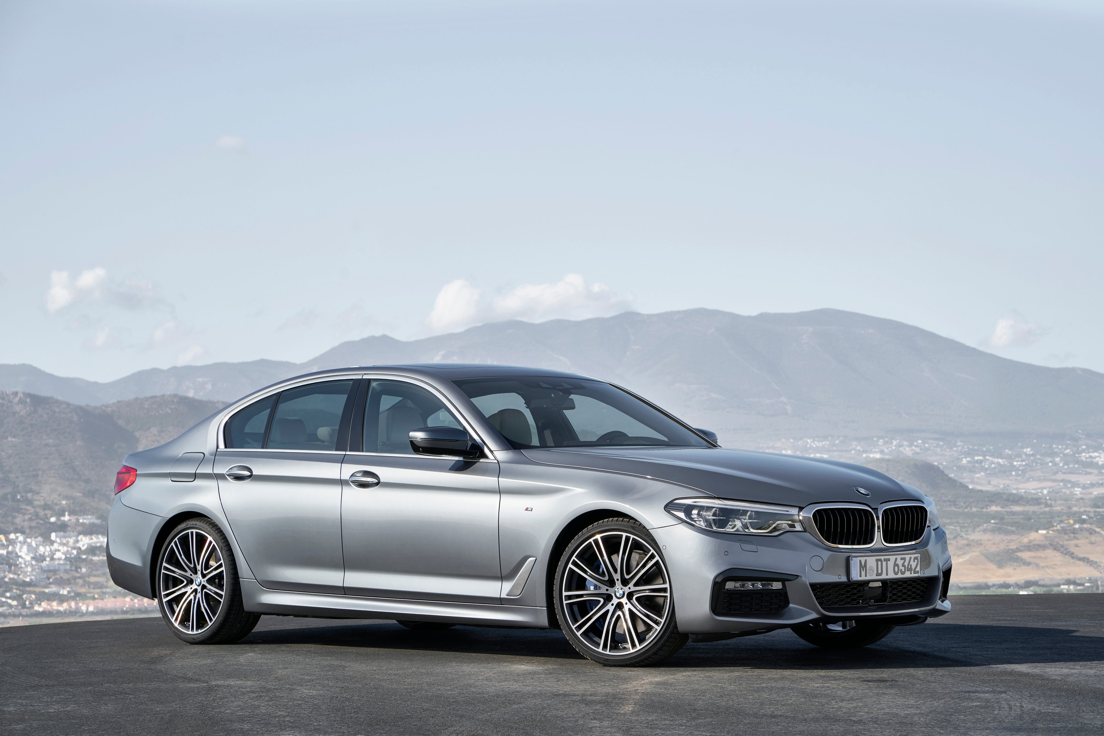 A silver BMW 5 Series sedan parked outdoors in bright sunlight, with mountains visible in the background.