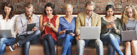 Men and women packed into a long couch, looking at laptops, tablets, and smartphones.