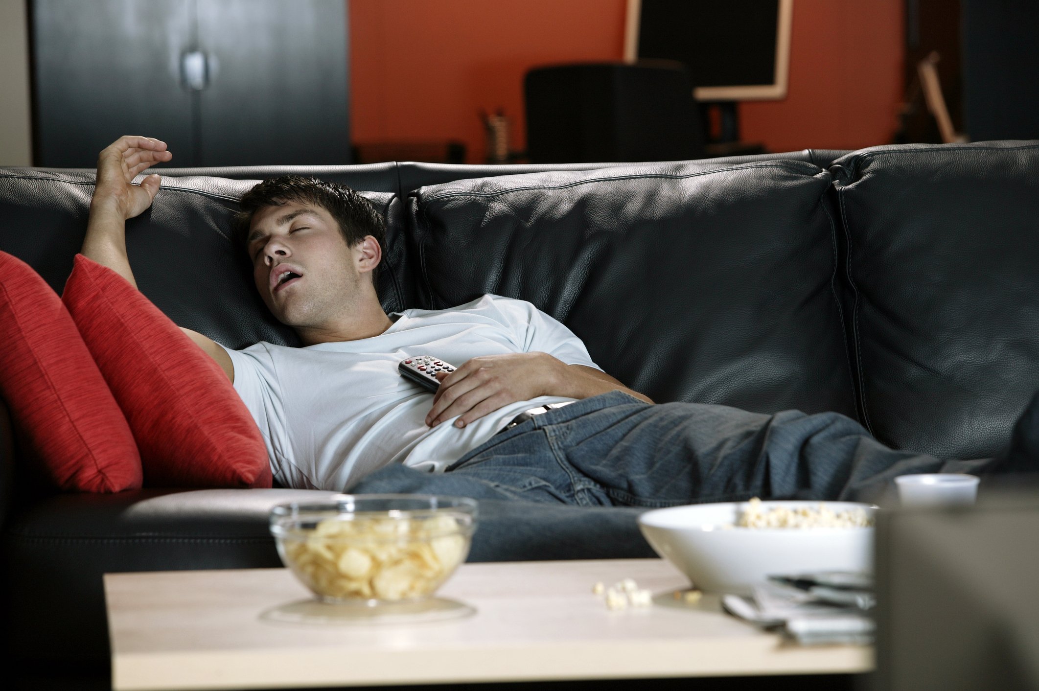 Young man sprawled on a couch with eyes closed and open mouth, holding a TV remote against his chest.