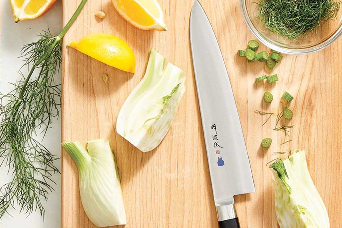 A chef's knife on a cutting board in the middle of fennel, lemon, and dill