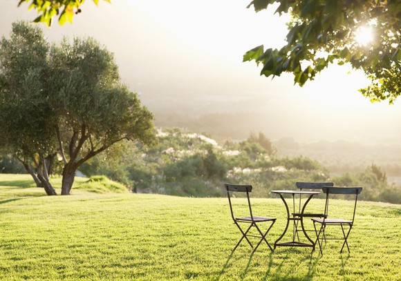 The sun shines through the leaves of a tree onto a green backyard and casts shadows on a round table and three chairs.