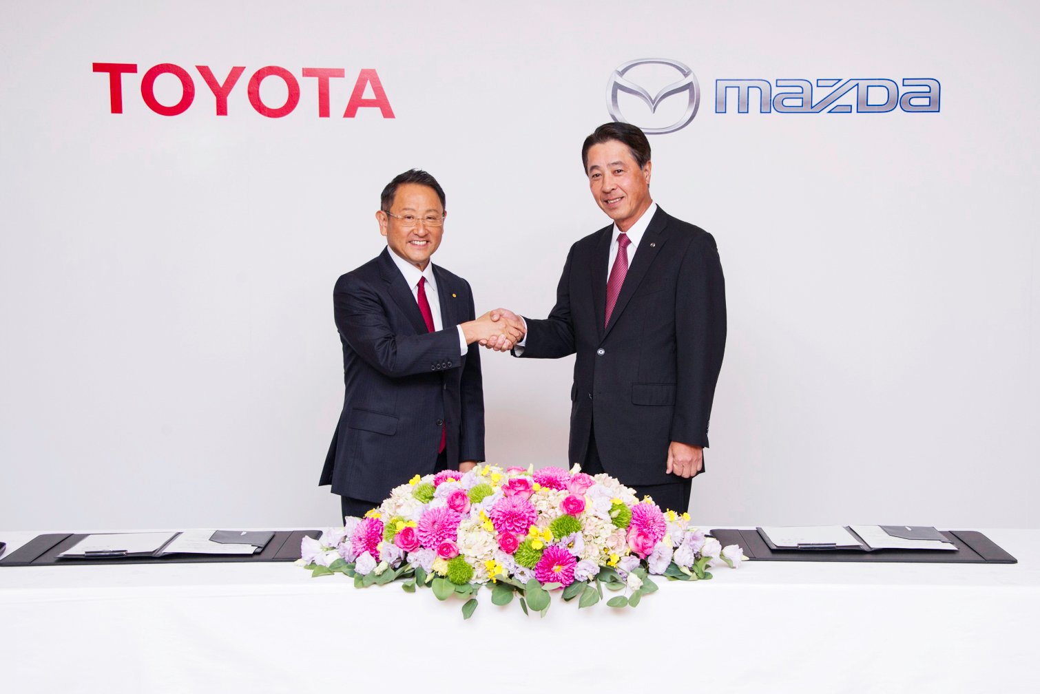 Akio Toyoda and Masamichi Kogai are shown shaking hands before a backdrop with the Toyota and Mazda logos. 