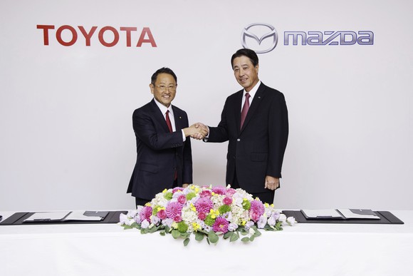 Akio Toyoda and Masamichi Kogai are shown shaking hands before a backdrop with the Toyota and Mazda logos. 