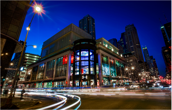 A lighted Under Amrour multi-story store in downtown Chicago on a corner at night, time laps photo shows lights blurred from cars traveling through the intersection in front of the store.