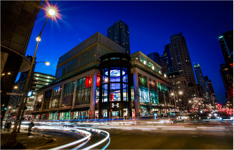 A lighted Under Armour's multi-story store in downtown Chicago at night with time lapse effects of car lights leaving a trail around the corner and across the busy street.