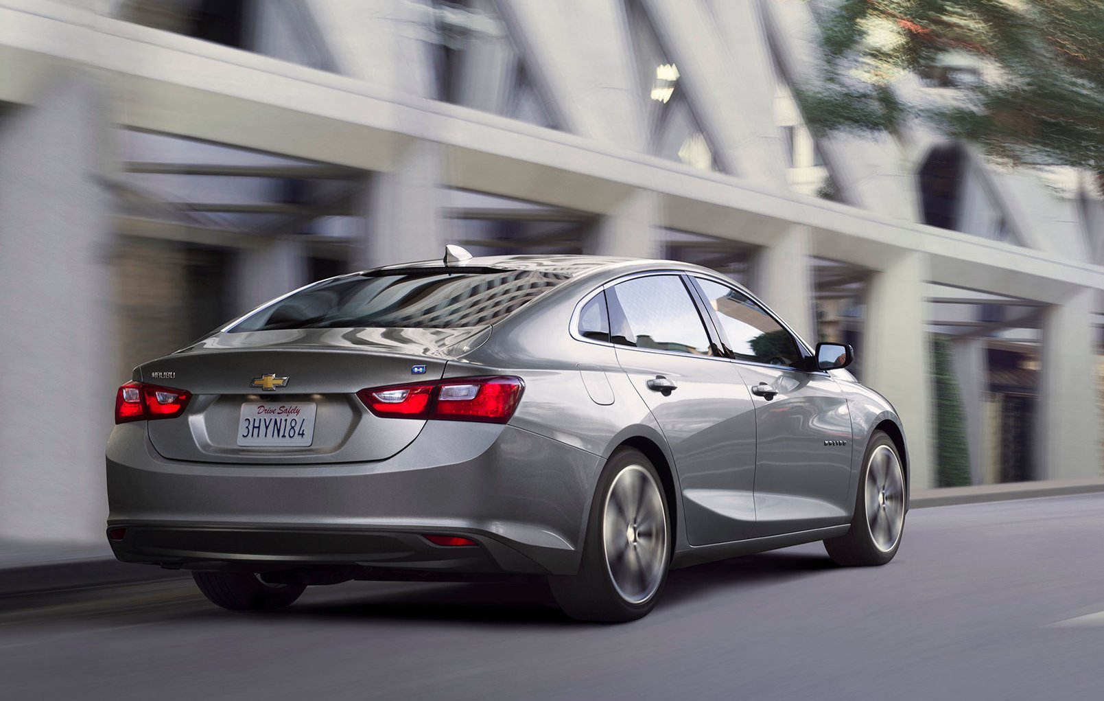 A 2017 Chevrolet Malibu Hybrid sedan in silver, driving past a white-framed commercial building.