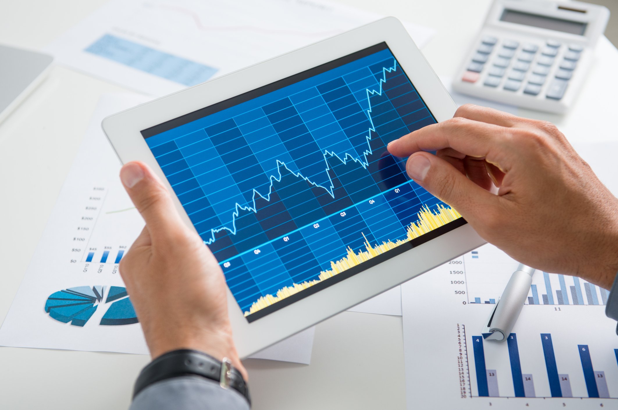 Hands holding a tablet computer showing a rising stock chart, above a table covered in other charts, pens, and a calculator
