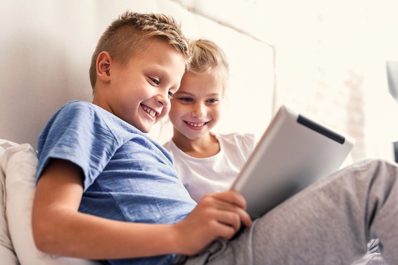 A young boy and girl on a bed, smiling at their tablet screen.