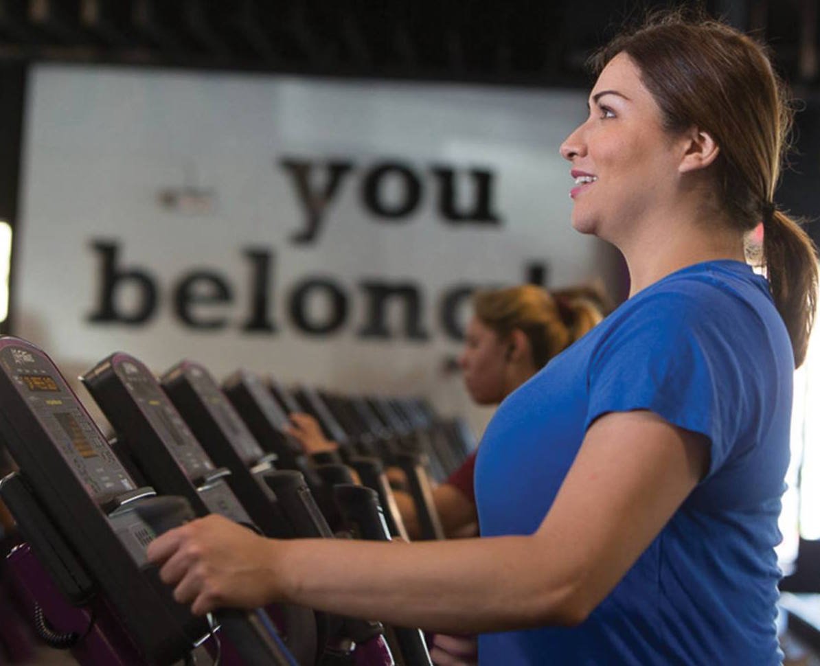 A woman on an exercise machine with a sign that says "you belong" on the wall behind her 