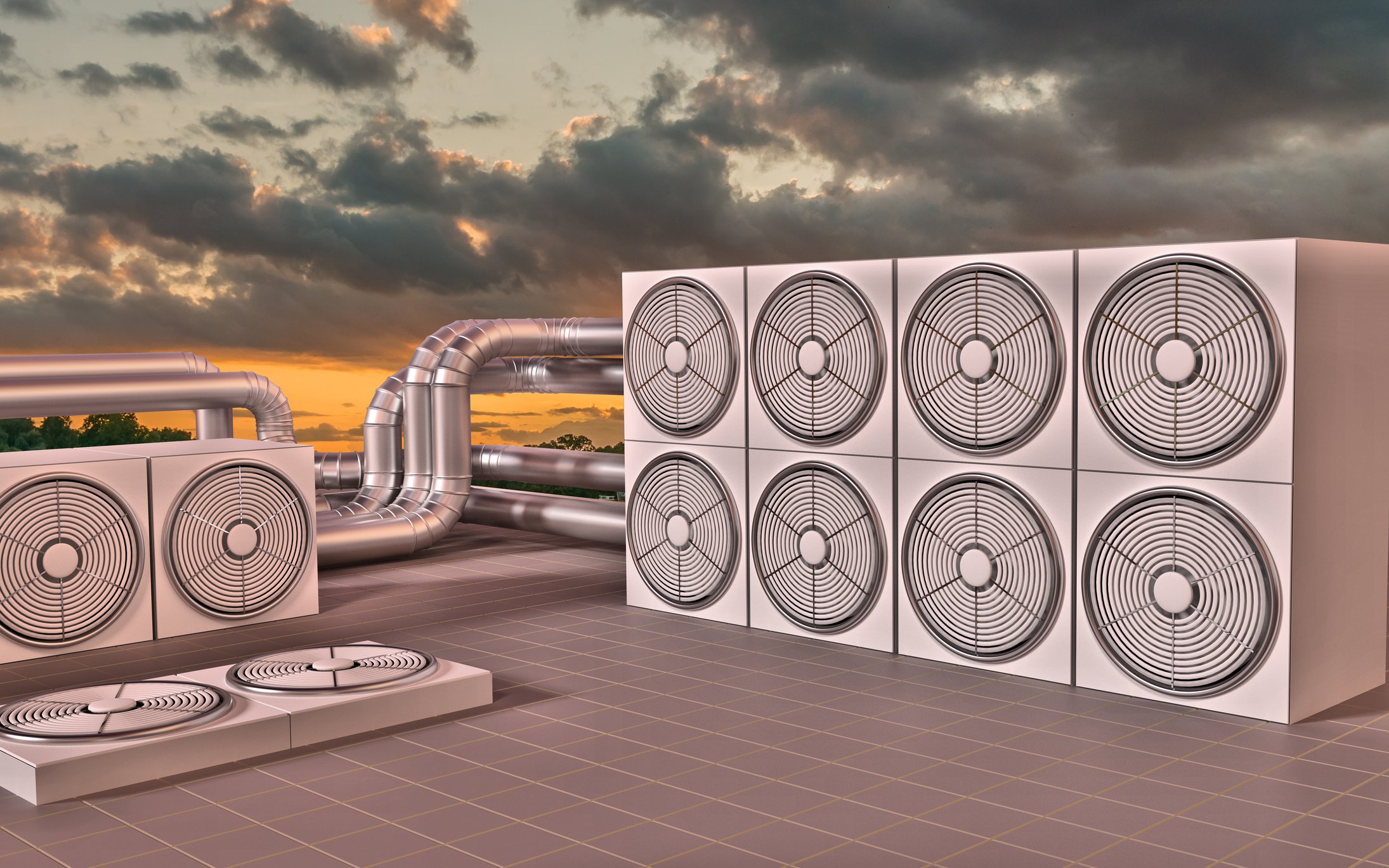 A cluster of industrial air conditioning units on the roof of a building, with a setting sun and cloudy sky in the background.