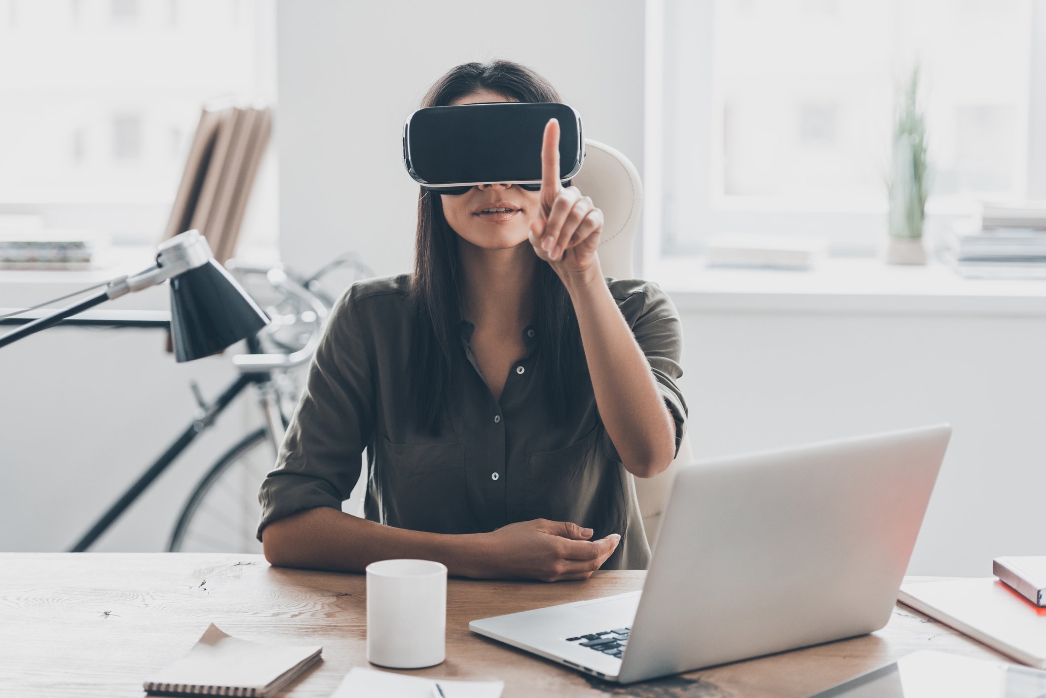 Woman sitting at desk wearing a virtual reality head and pointing her finger in the air.