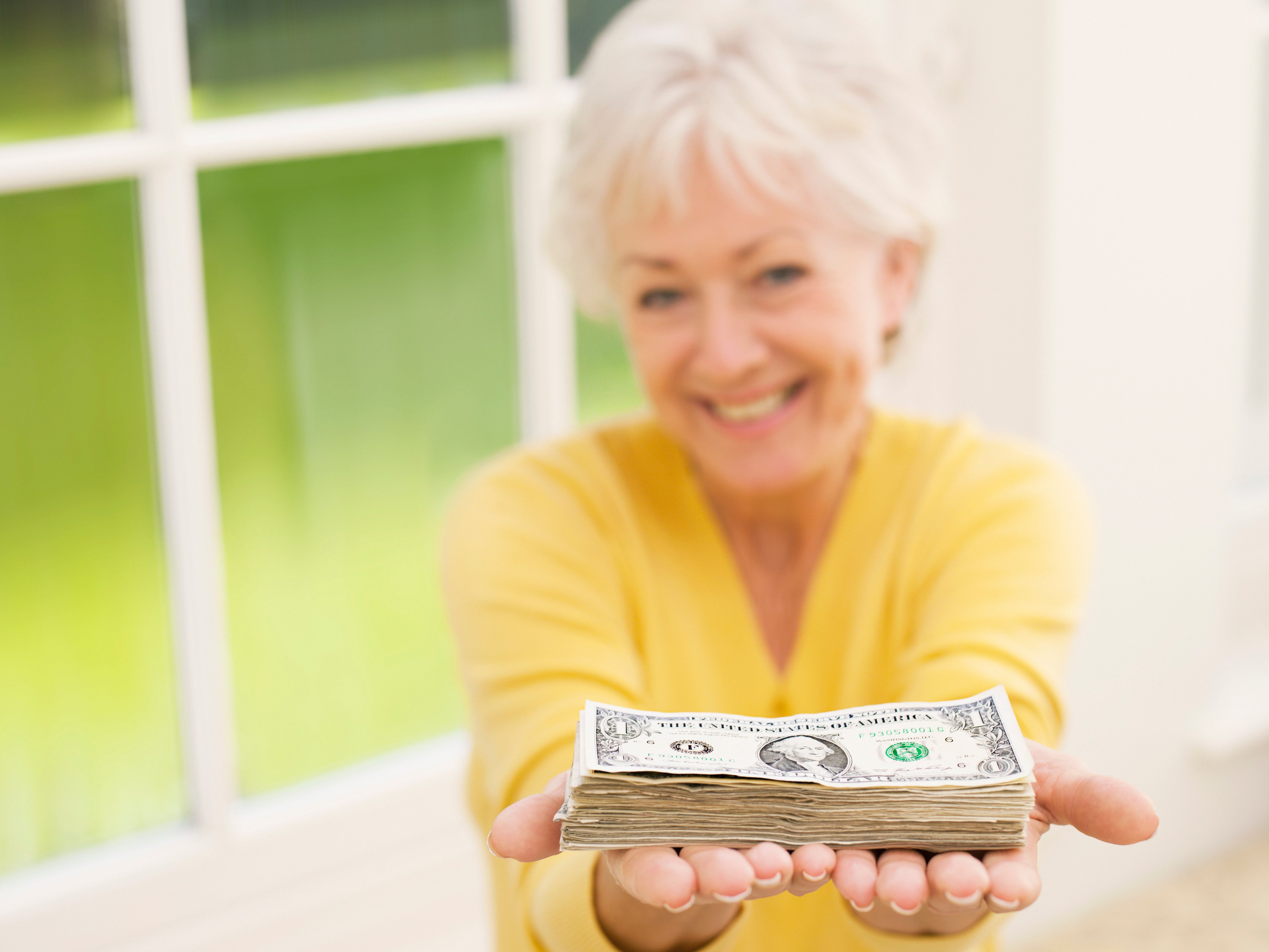 An old woman wearing a yellow shirt holding out a pile of money and smiling.