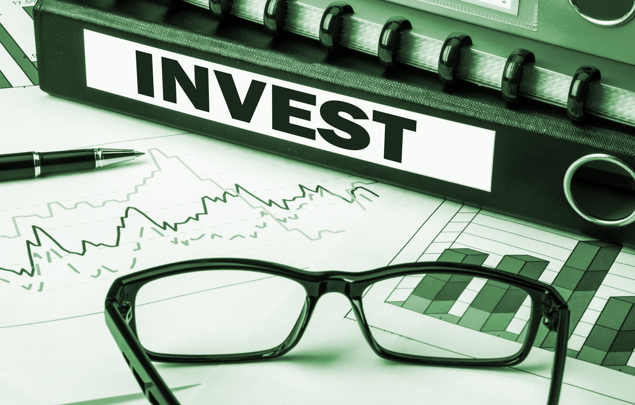 A binder labeled "invest" sits on top of a stock chart and next to a pen and pair of glasses.