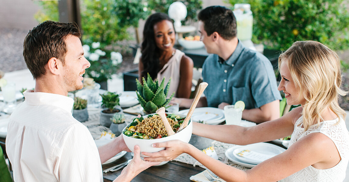 A man passes a bowl of noodles to a woman as a group is sat around a dinner table.