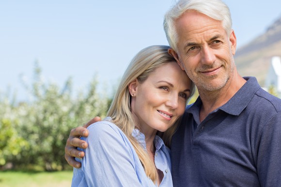 An older couple stands smiling outdoors, with the man's arm around the woman's shoulder.