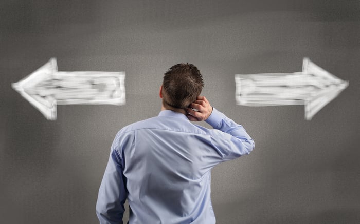 A man in a blue business shirt stands with his back to the viewer, scratching his head over two white arrows on the wall in from of him pointing in opposite directions.