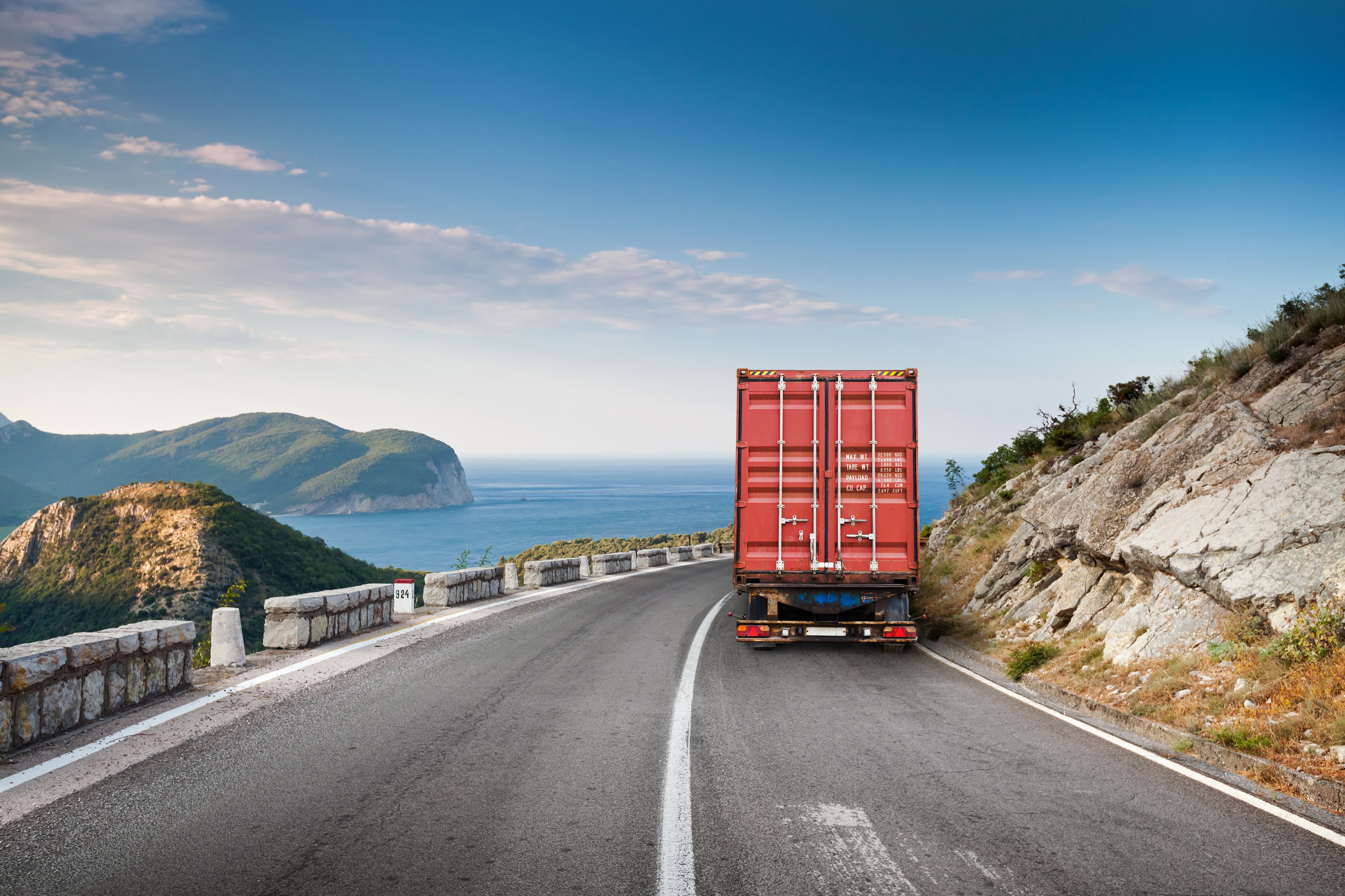 View of the back of a red truck traveling on a two-lane highway with mountain on one side and a body of water on other side. 