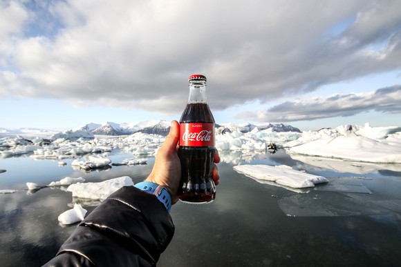 A hand holds a bottle of Coca-Cola backed up by a glacier vista.
