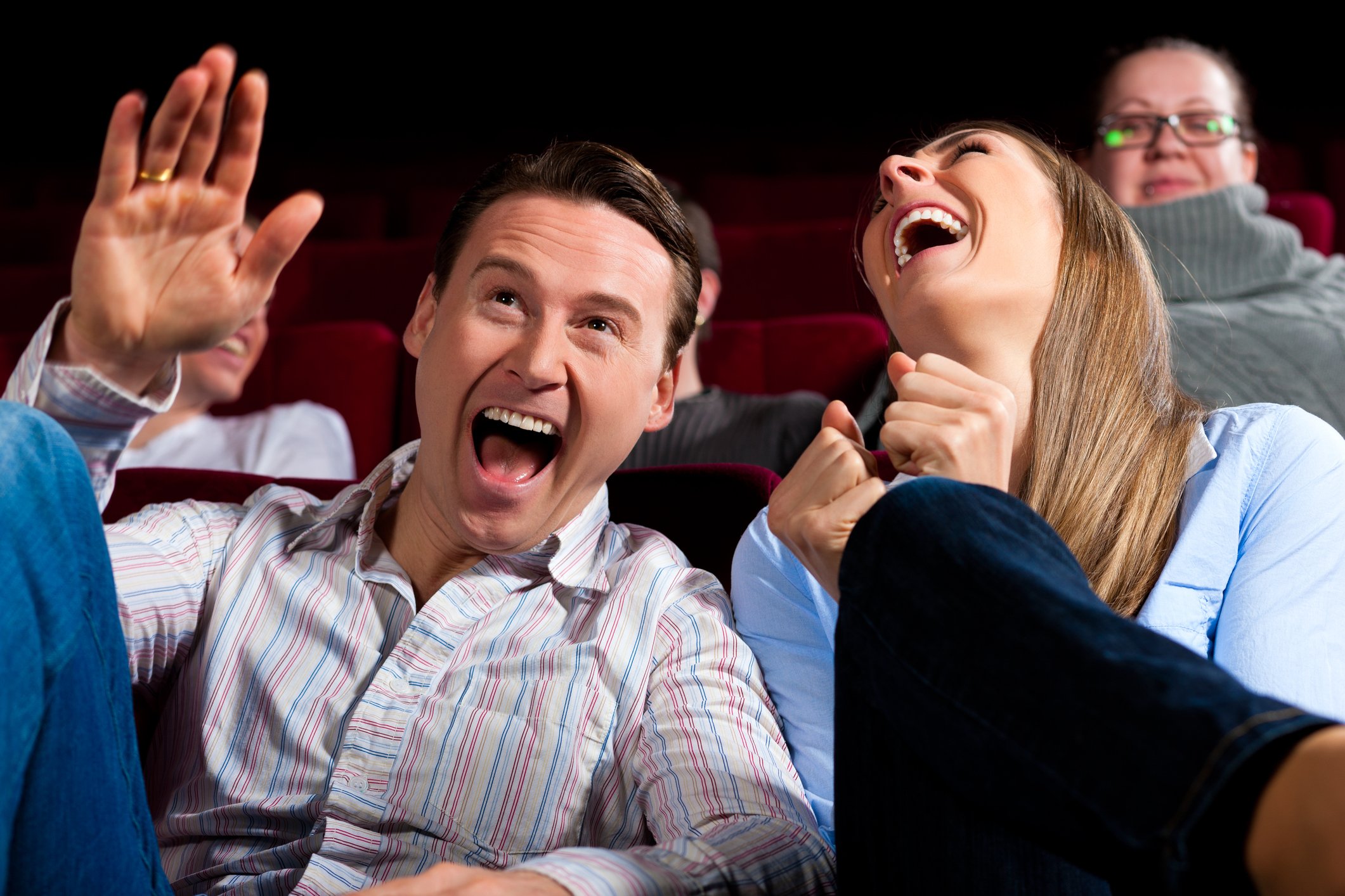 Middle-aged man and woman sitting in a theater and laughing hard, suggesting they're watching a funny scene in a movie. A few other people are visible in seats behind them.
