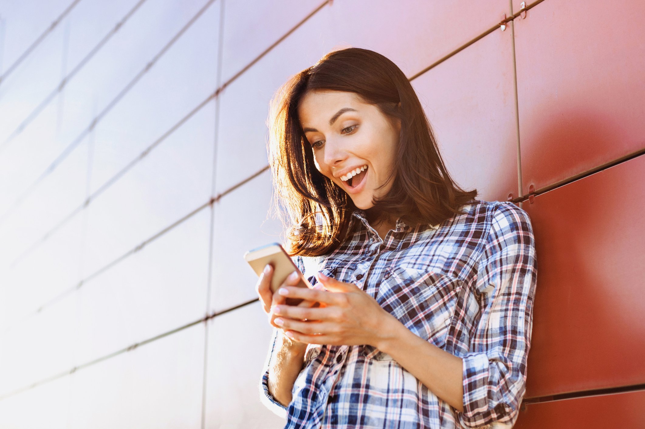 A woman smiles down at her smartphone as she leans against a wall.