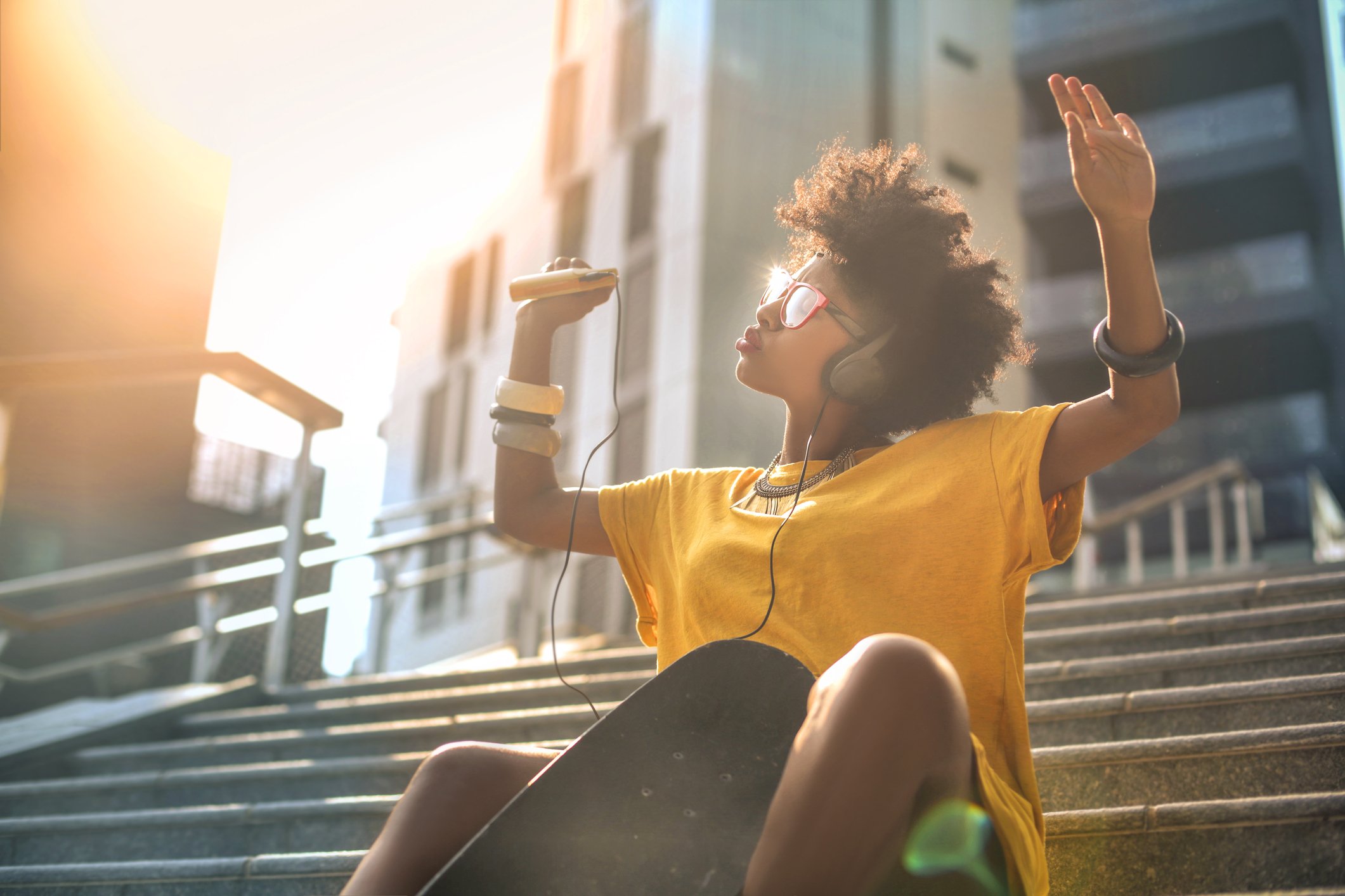 A woman in a yellow shirt sits outside on a flight of steps, raising her arms in the air as she listens to music.
