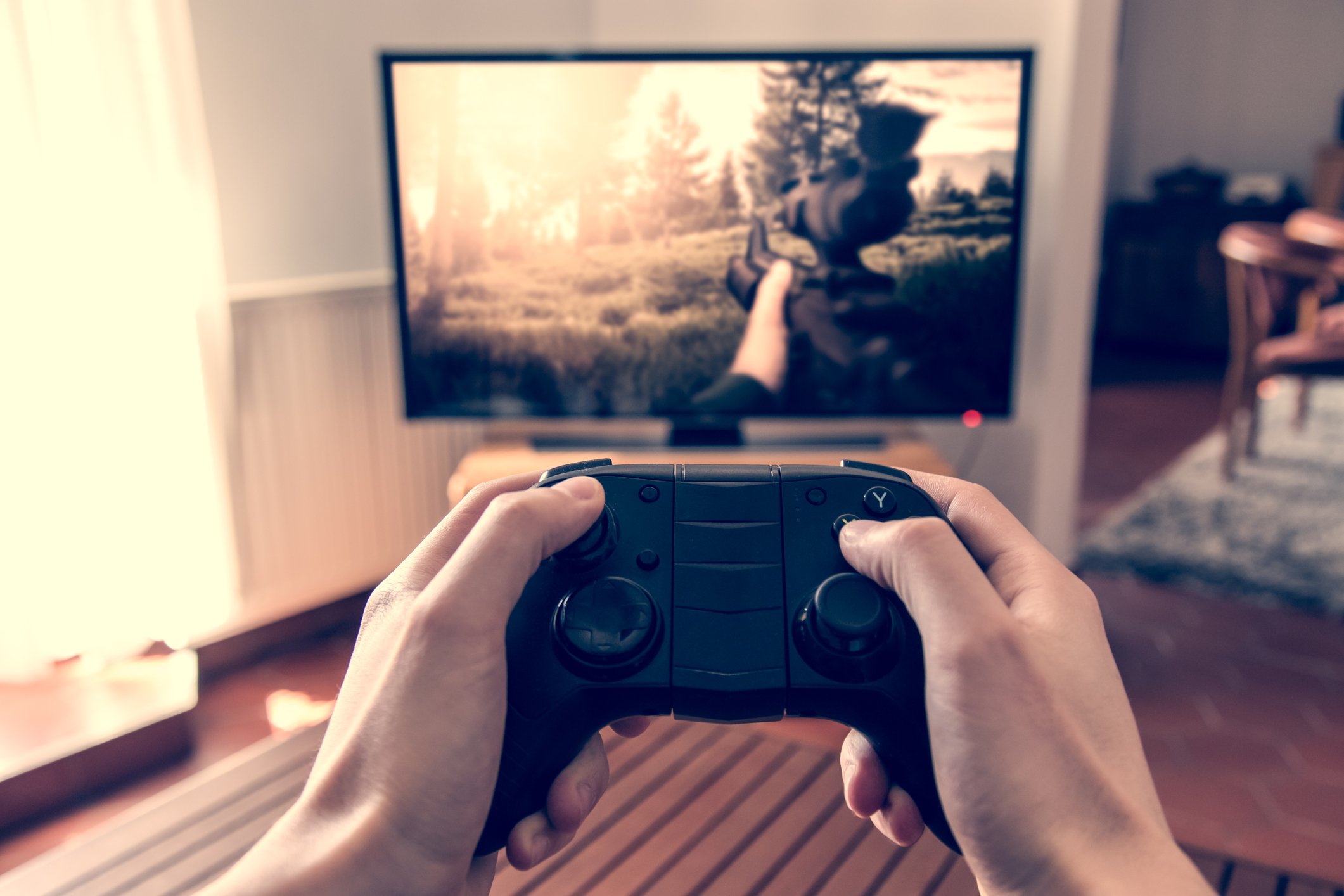 Man's hands holding a video game controller with a large TV screen displaying a game in the background.