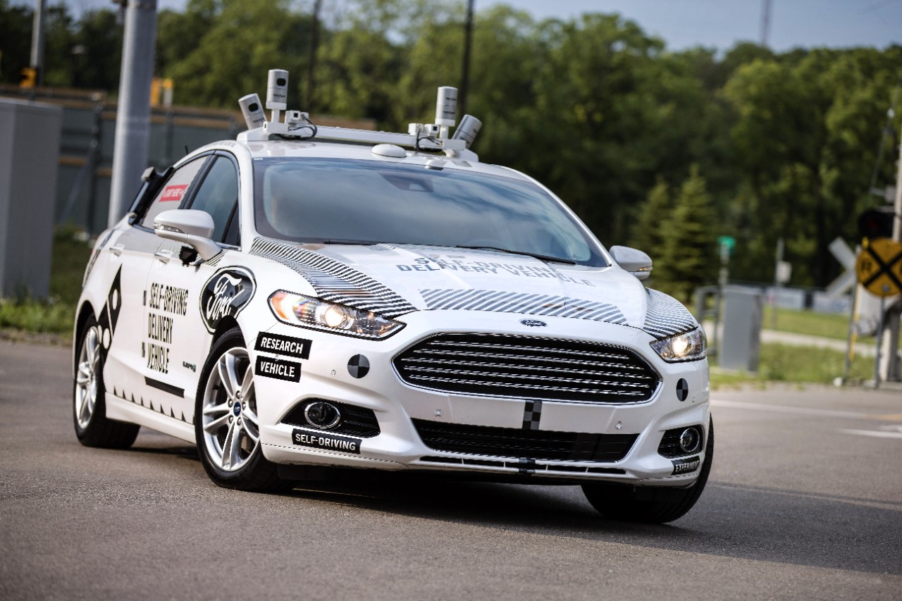 A white Ford Fusion Hybrid with self-driving sensors and logos indicating that it's a self-driving Domino's Pizza delivery vehicle. 