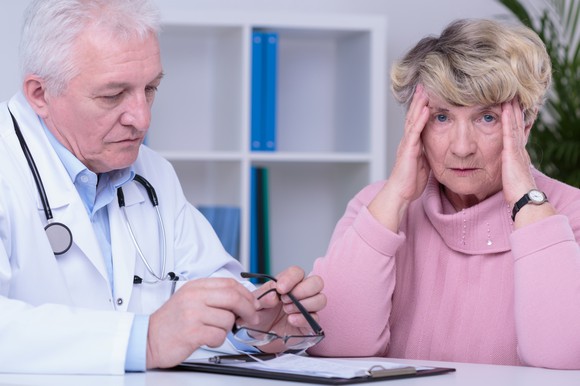 A senior woman holding her head in frustration while having a discussion with a doctor. 