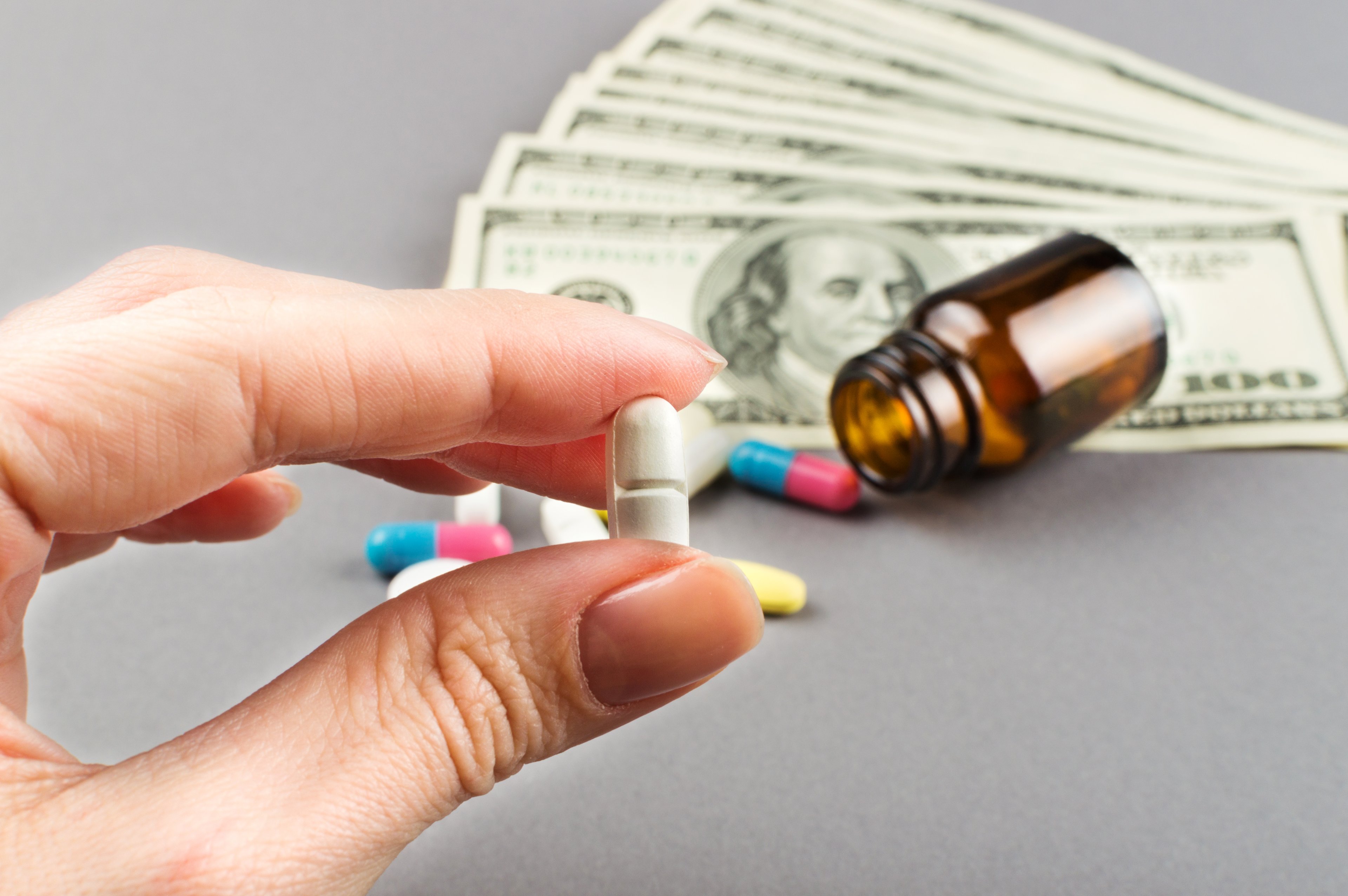 Woman's hand holding pill with money, pill bottle, and pills in background