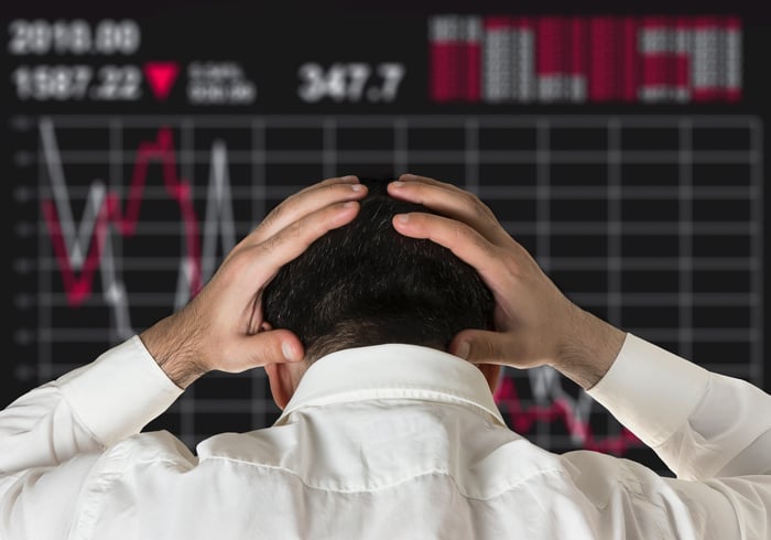 A man holds his head in his hands as he watches a screen showing a declining stock price.