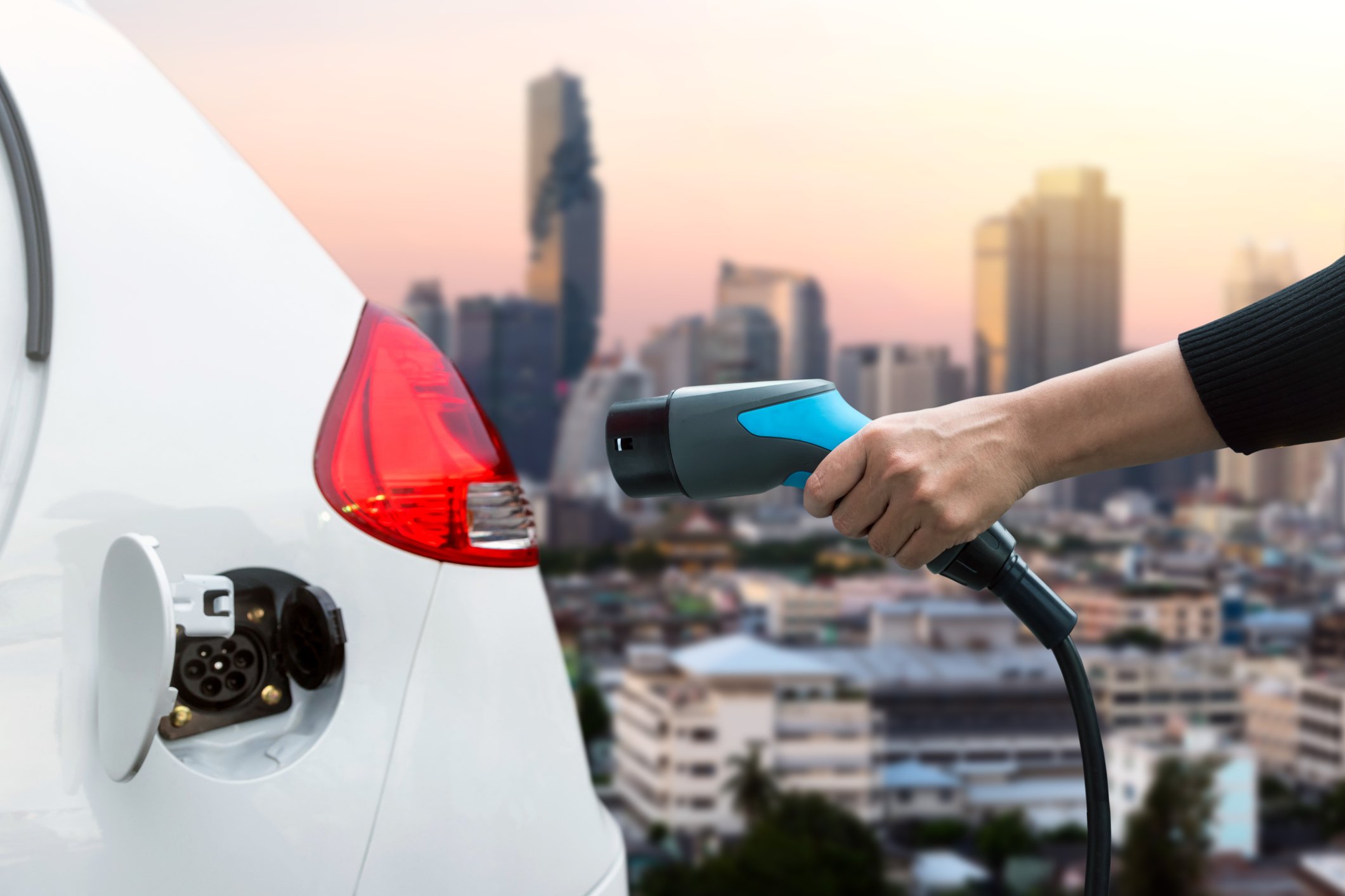 Woman's arm plugging in an electric vehicle with a city skyline in the background.