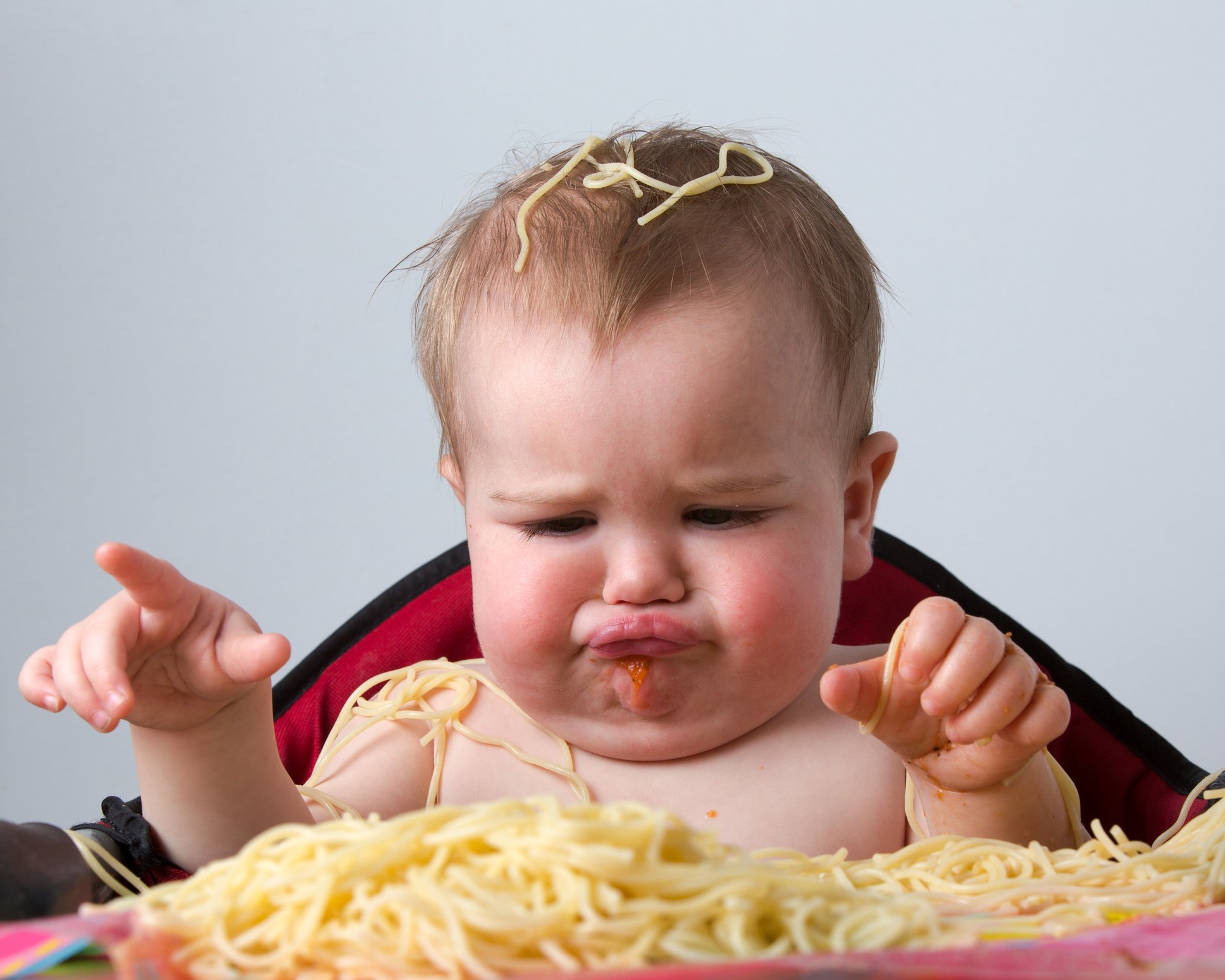 Frowning baby sitting in high chair eating spaghetti noodles with hands and making a mess.