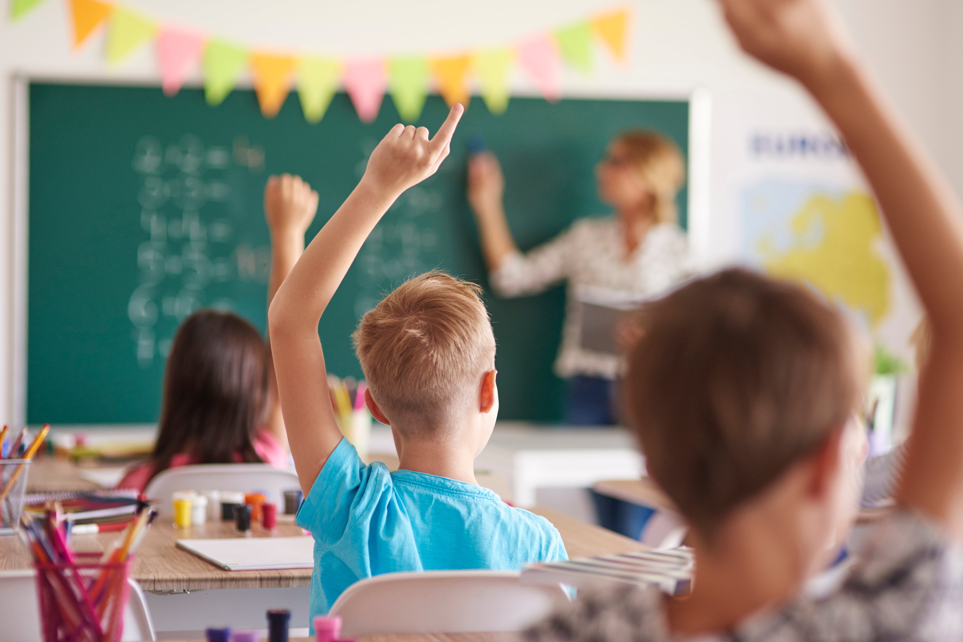 Children raise their hands in a classroom as a teacher in the background points to the chalkboard.