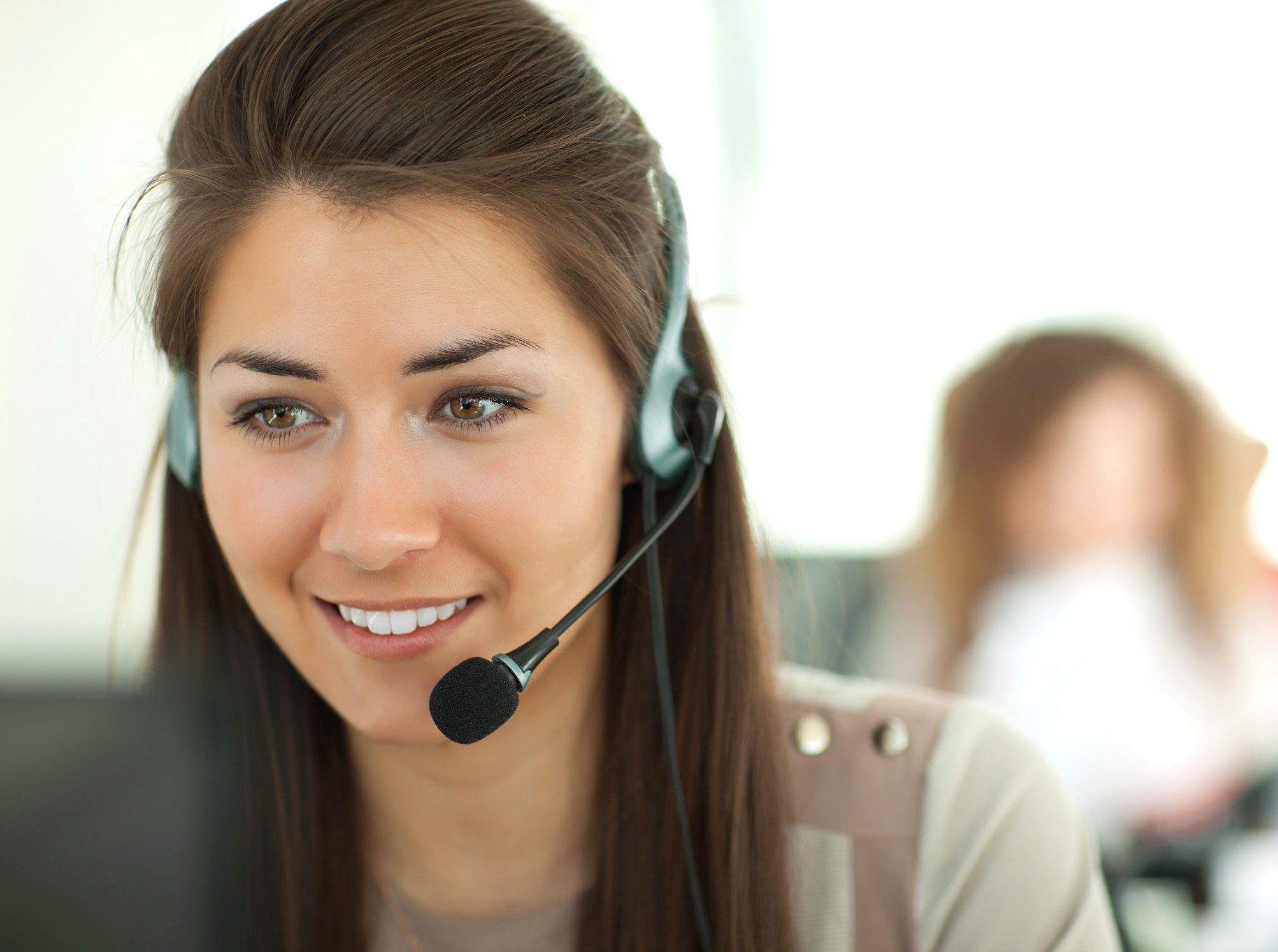 A smiling woman in an office setting wears a telephone headset.
