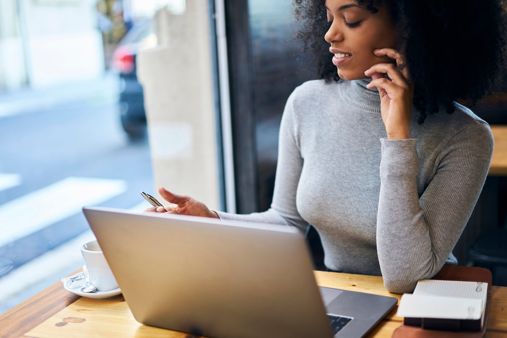 A woman sits at a laptop near a window, with a coffee cup next to her.