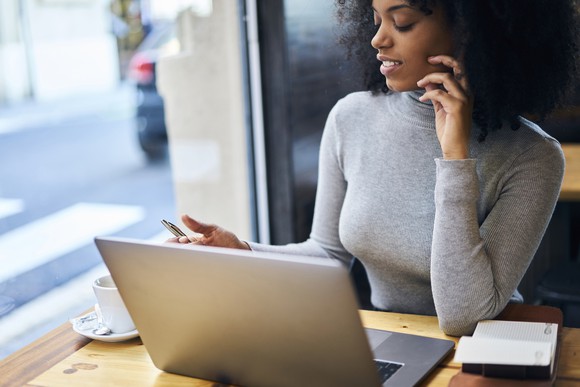 A woman sits at a laptop near a window, with a coffee cup next to her.