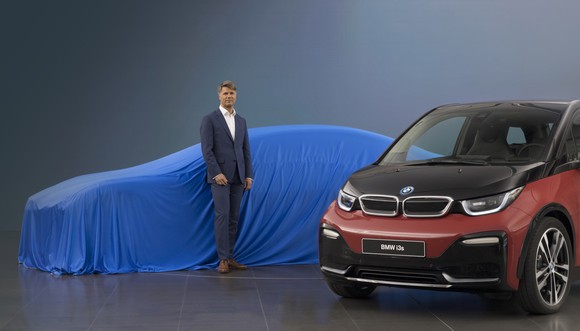 Krueger is standing in front of a low-slung car covered with a blue cloth. A BMW i3 hatchback is parked next to him. 