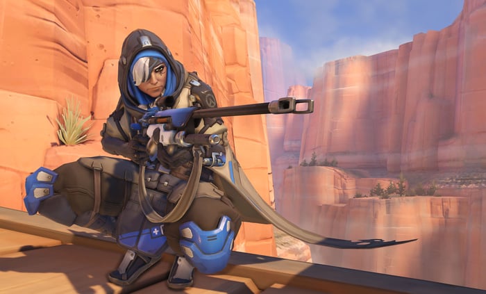 "Overwatch" character in a desert landscape, wearing a hood and holding a sniper rifle.