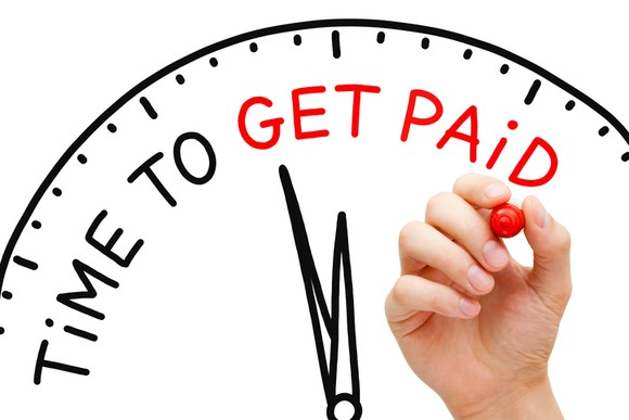 Hand writing the words "Time To Get Paid" with red marker on the drawing of a clock on a whiteboard.