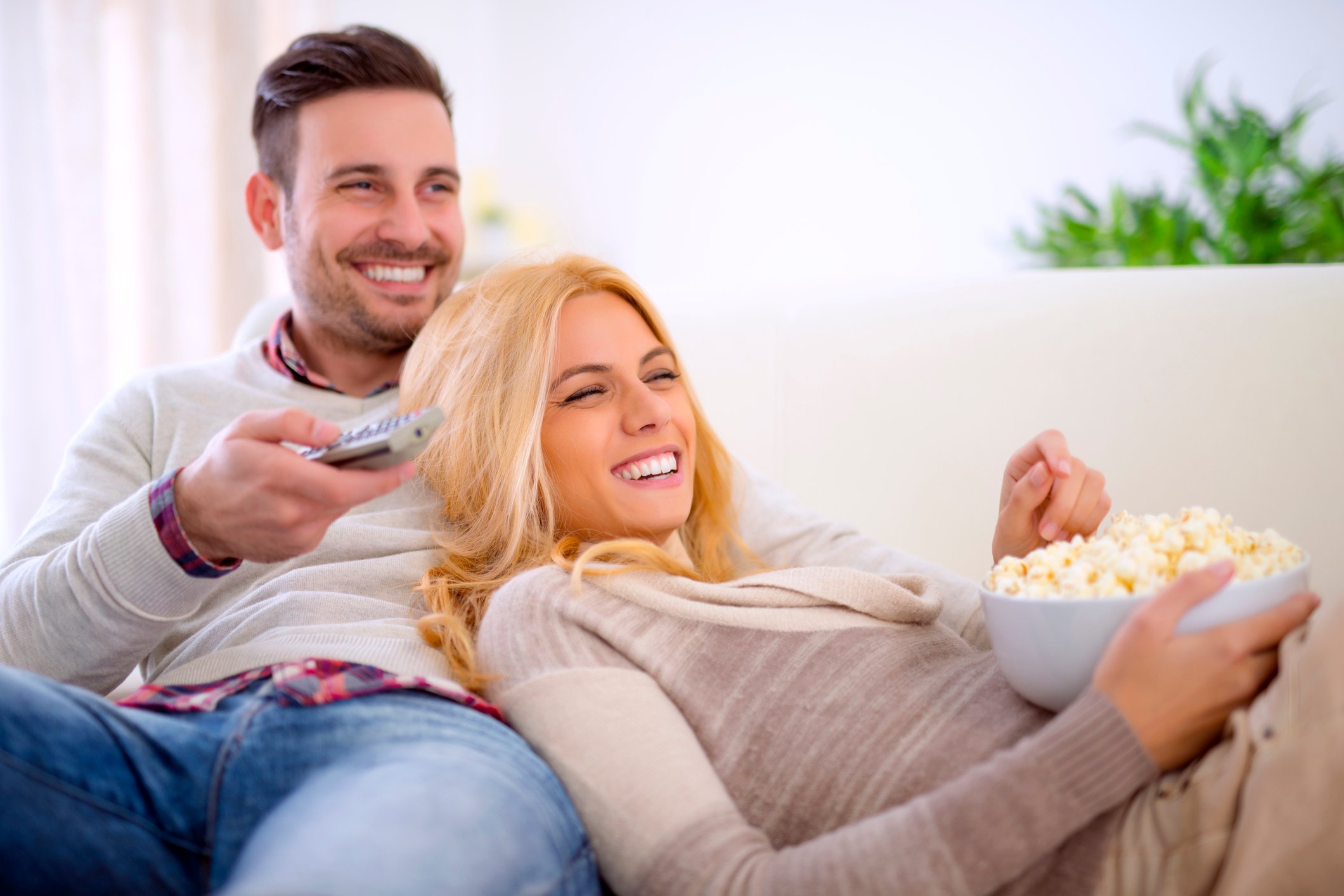 A smiling couple relaxing on a couch, with popcorn and TV remotes close at hand.