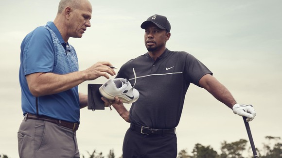 Tiger Woods leans on a golf club and talks with a man holding a Nike shoe.