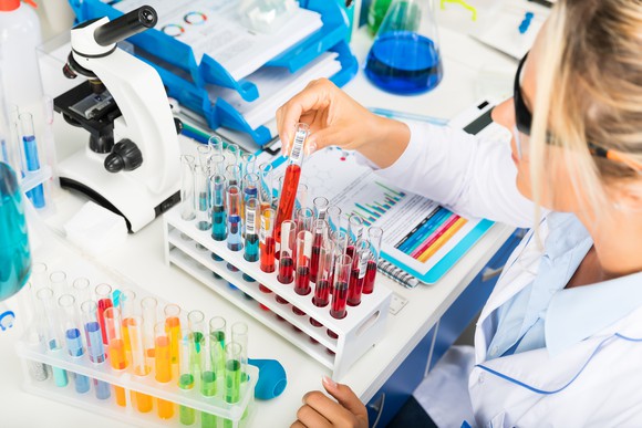 A seated scientist with test tubes and a microscope on table in front of her.