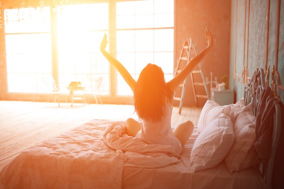 With sun pouring in from a window across the room, a woman with her back to the camera sits up in bed with her arms raised, as if stretching.