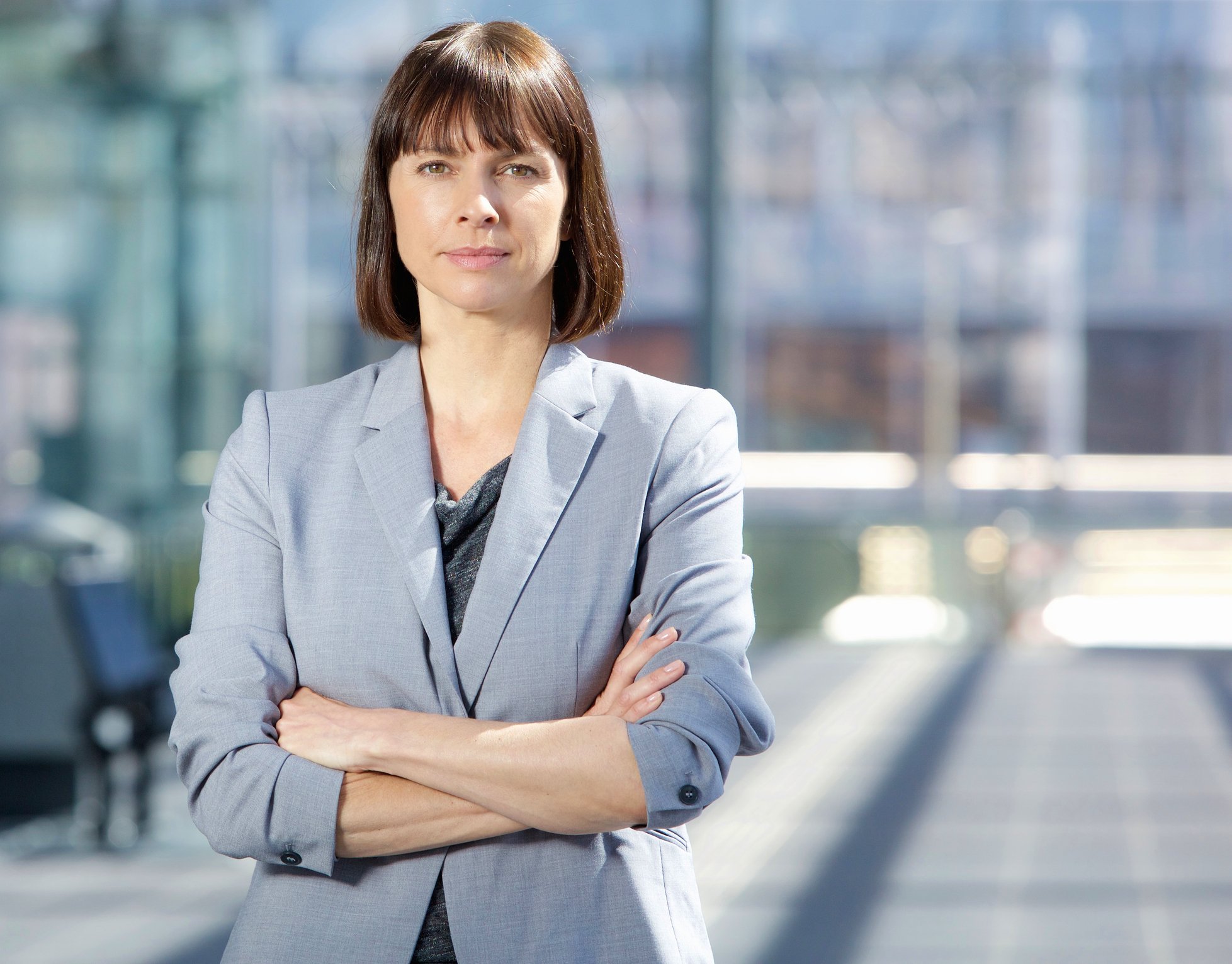 A woman in a light blue blazer stands outdoors with her arms crossed.