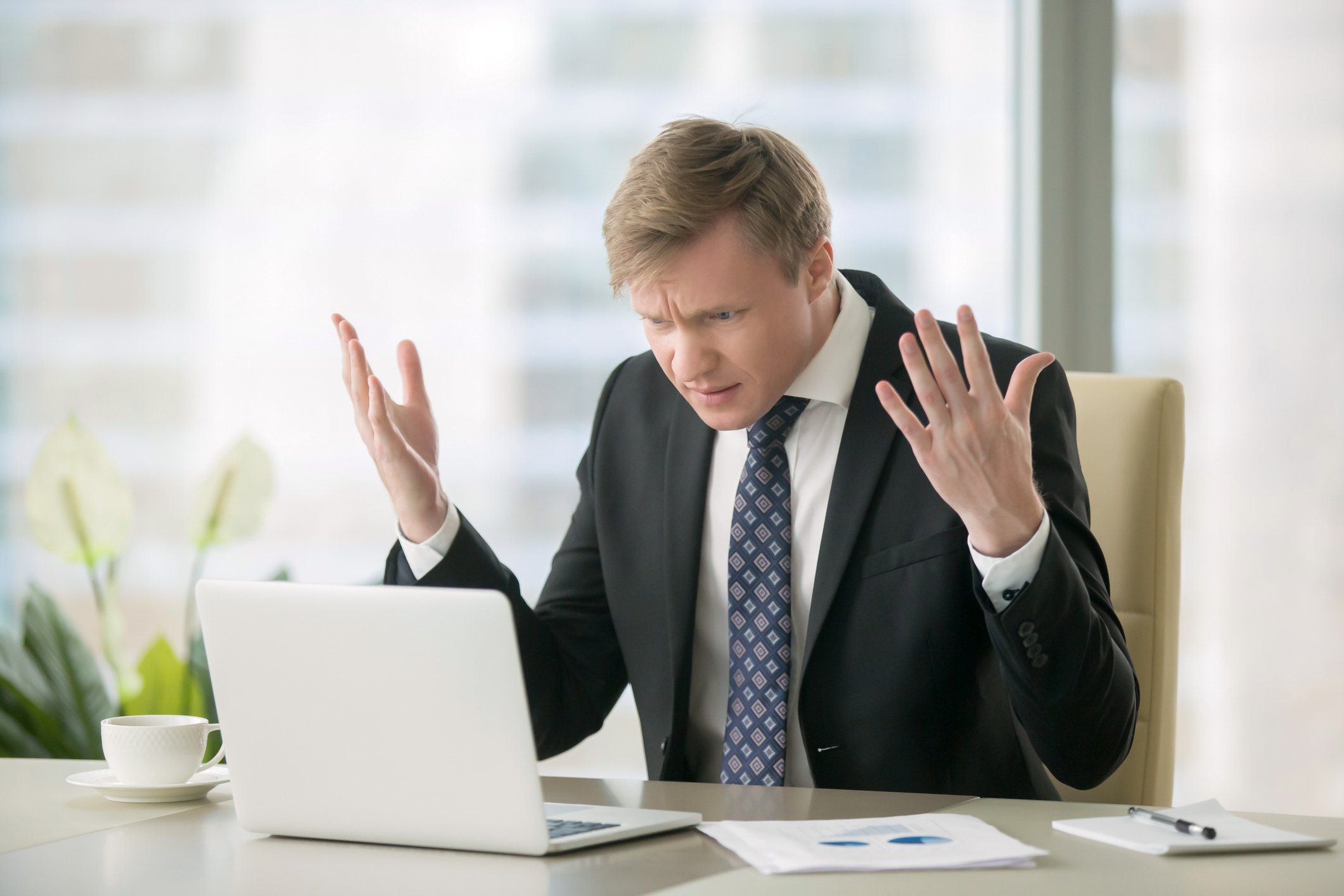 A man in a suit, sitting at a desk, looks at a computer with both hands up in the air in frustration.