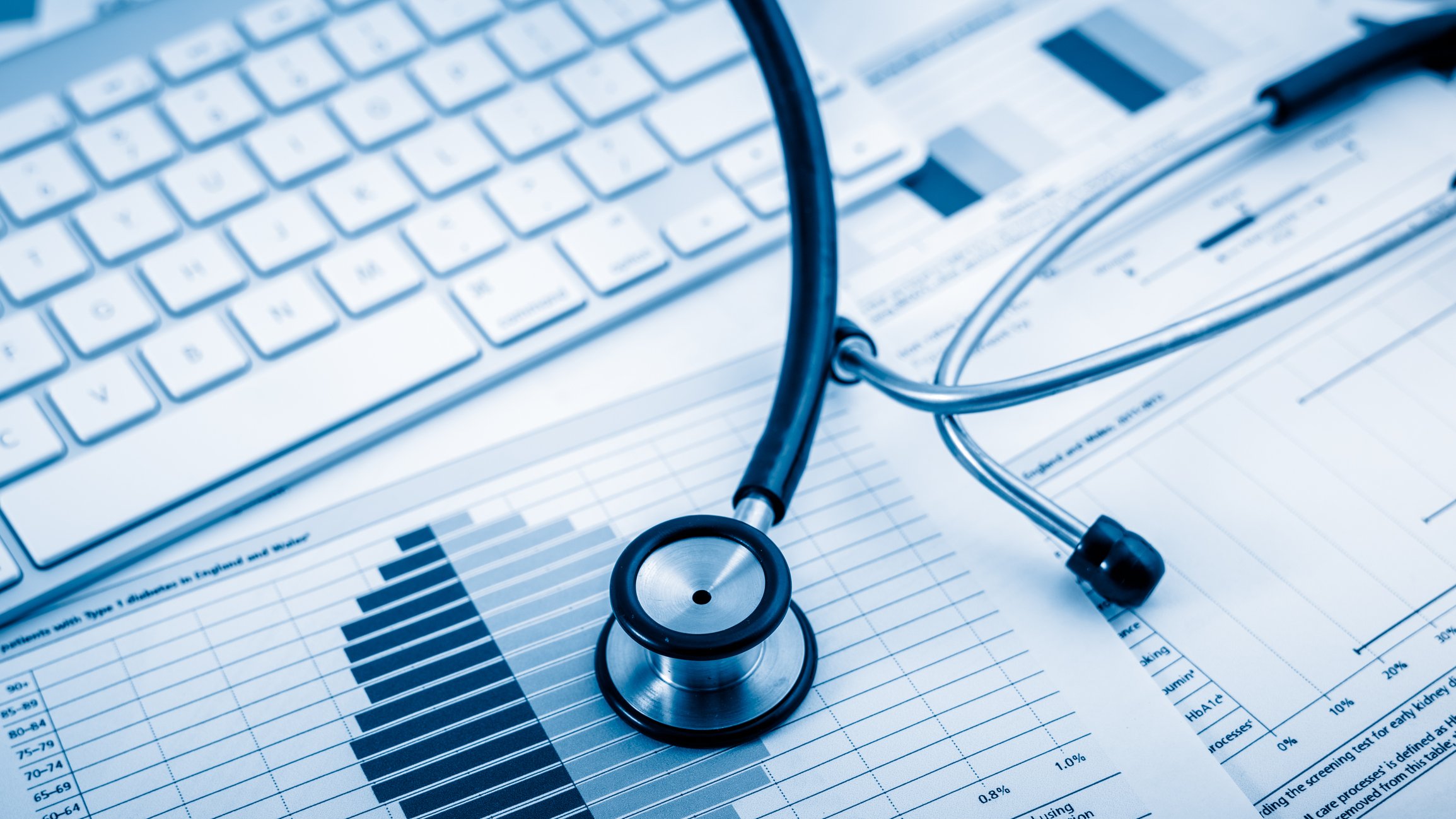 A stethoscope sits on top of financial statements, which are spread out next to a computer keyboard.