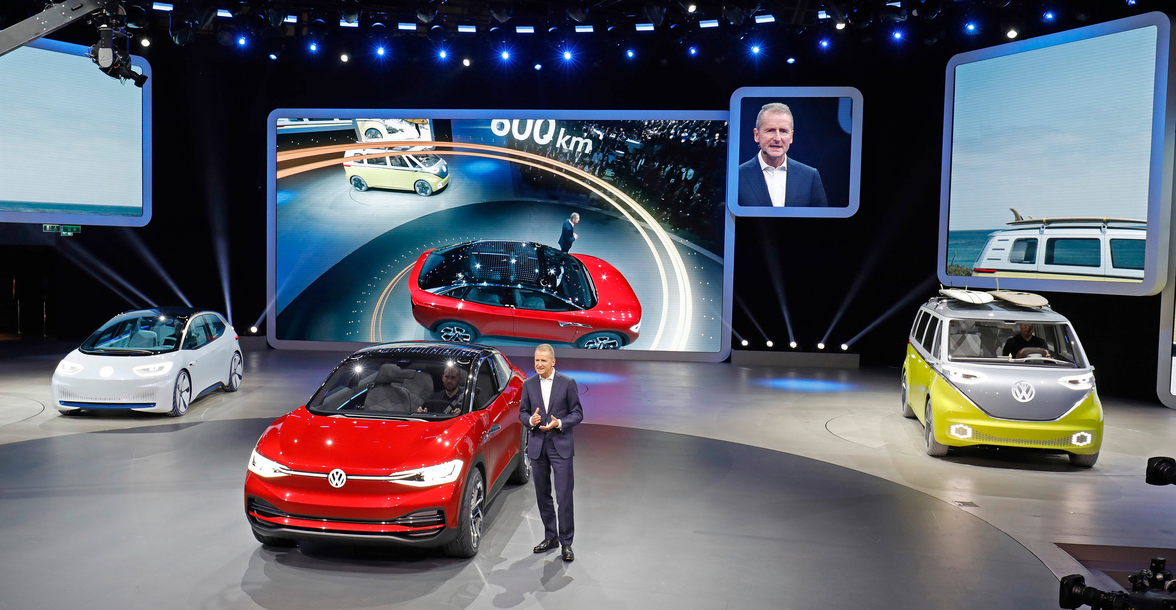 Diess is standing next to the red I.D. Crozz II on an auto-show stand. The white I.D. hatchback is to the left, and the yellow and silver I.D. Buzz is at the right. 