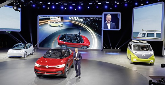 Diess is standing next to the red I.D. Crozz II on an auto-show stand. The white I.D. hatchback is to the left, and the yellow and silver I.D. Buzz is at the right. 
