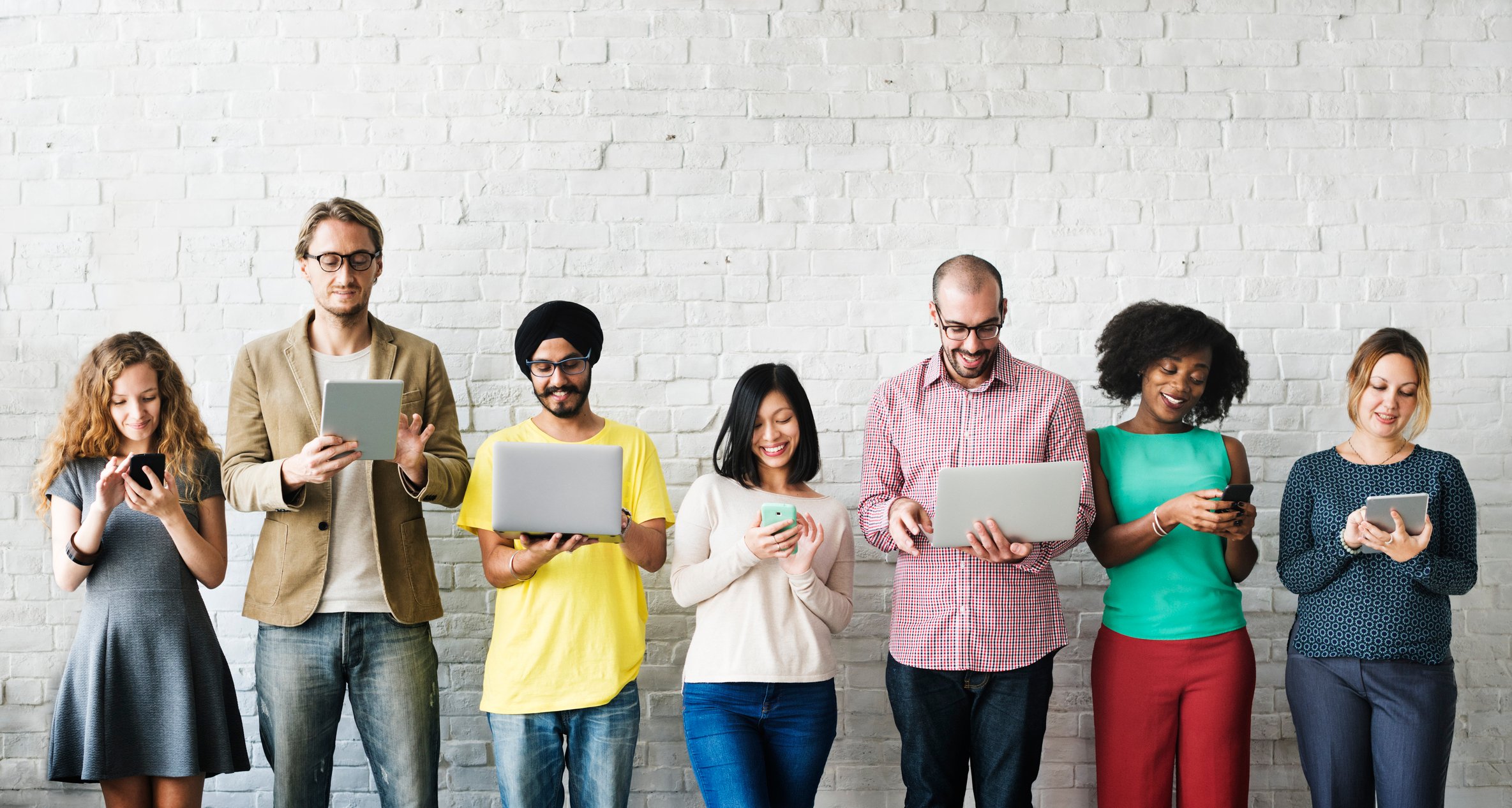 A group of 7 people from various cultures smiling at their smartphones, laptops, and tablets.