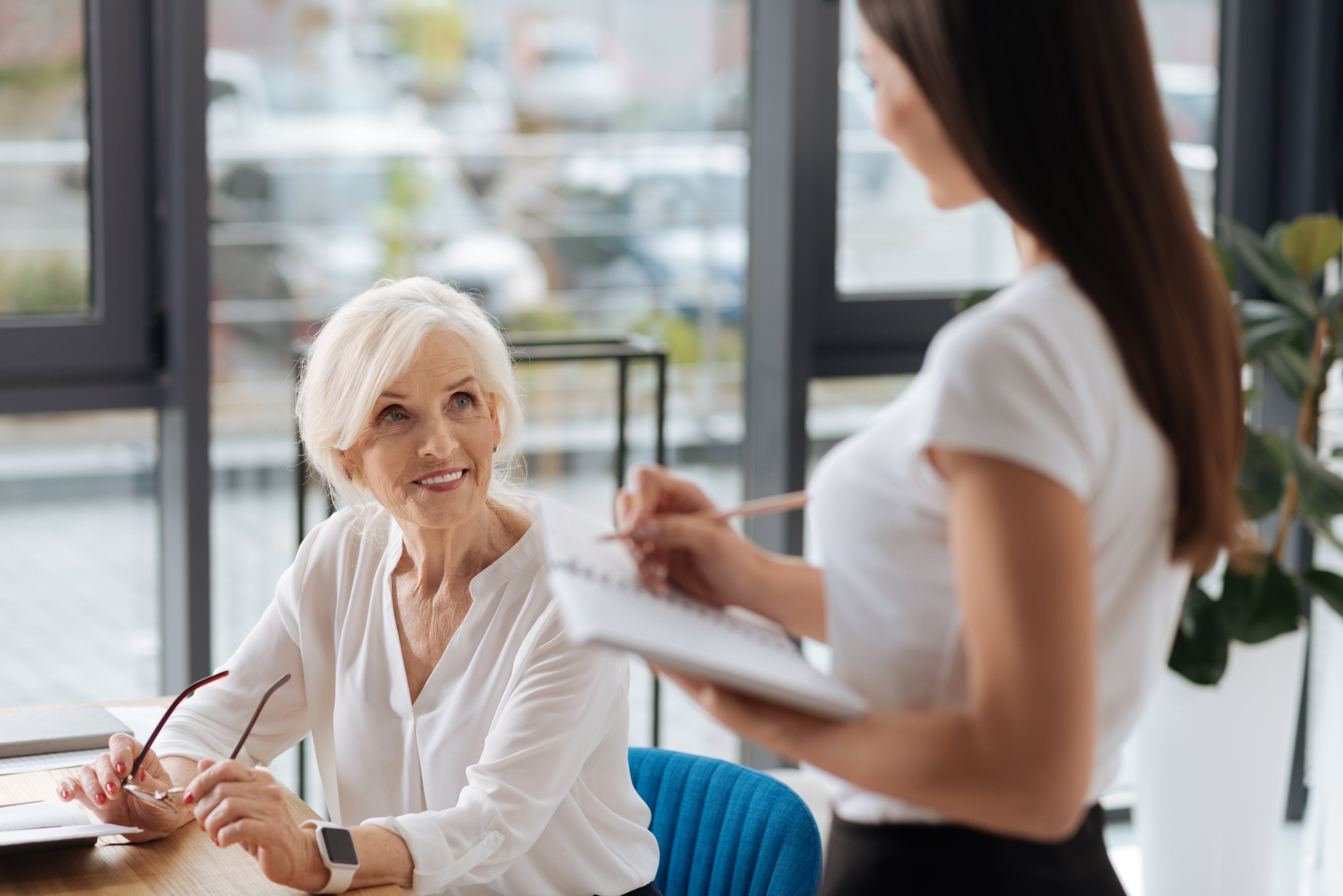 Woman sitting at table, talking to standing woman holding a notebook and writing implement.